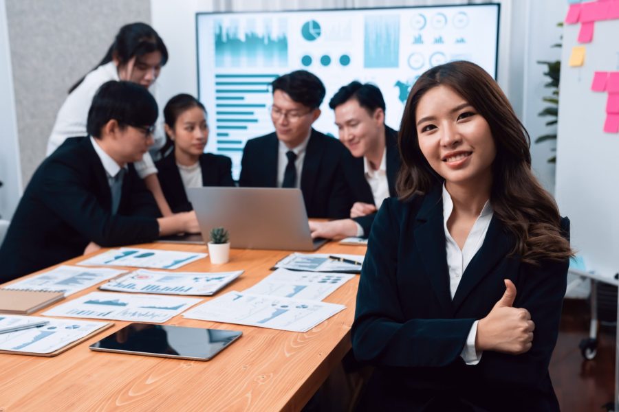 Focus portrait of female manger, businesswoman in the harmony meeting room with blurred of colleagues working together, analyzing financial paper report and dashboard data on screen in background.